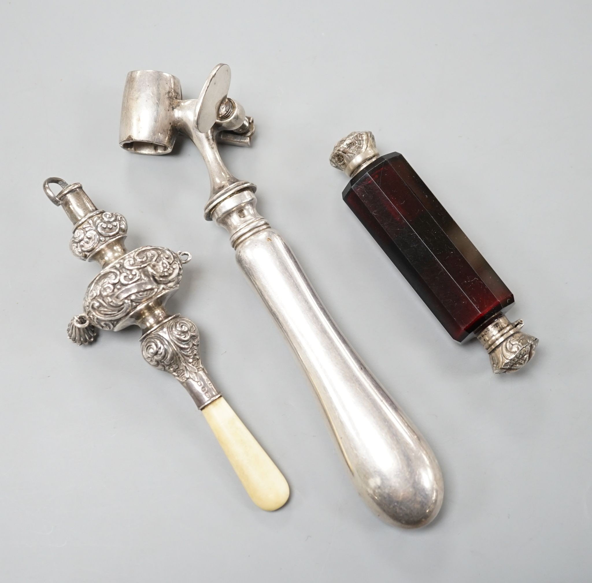 A silver mounted rattle, a white metal mounted double ended ruby glass cent bottle and a silver plated handled roast joint holder.